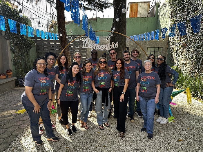 Group photo of a diverse team smiling outdoors in front of a 'Bienvenidos' sign, all wearing matching t-shirts that read 'Lighten the Load, Ease the Liftn