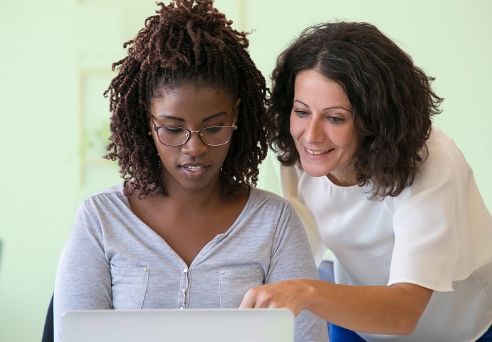 Two women side by side, one is writing in the notebook and the other is pointing at something on the notebook screen
