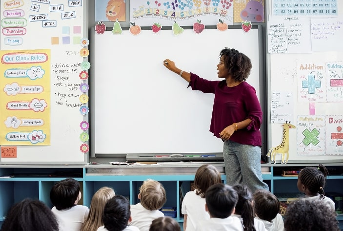 Teacher talking and writing on the board while the children sitting on the floor are paying attention