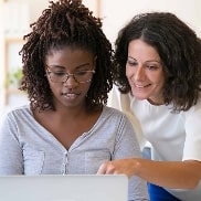 Two women side by side, one is writing in the notebook and the other is pointing at something on the notebook screen
